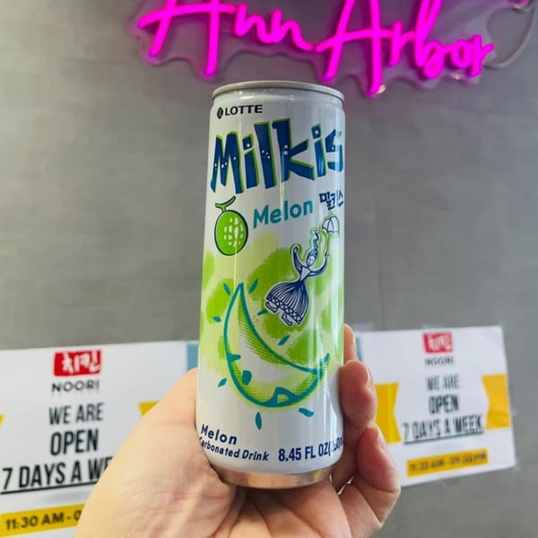 Hand holding a can of Lotte Milkis Melon carbonated drink, with a neon "Ann Arbor" sign in the background.