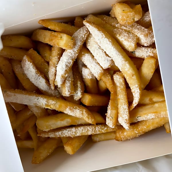 Close-up of golden french fries dusted with white powder, served in a white takeout box.