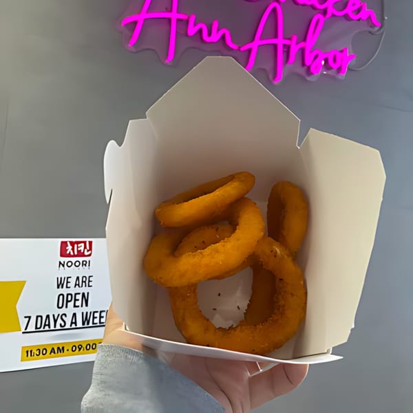 Hand holding a white takeout box filled with golden-brown onion rings from Noori Chicken Ann Arbor.