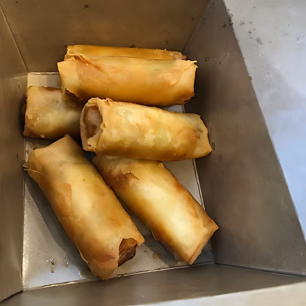 Six golden-brown egg rolls, likely from Noori Chicken, are nestled inside a takeout container.