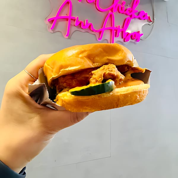 Hand holding a Noori Chicken sandwich with pickles and sauce, in front of a neon sign.